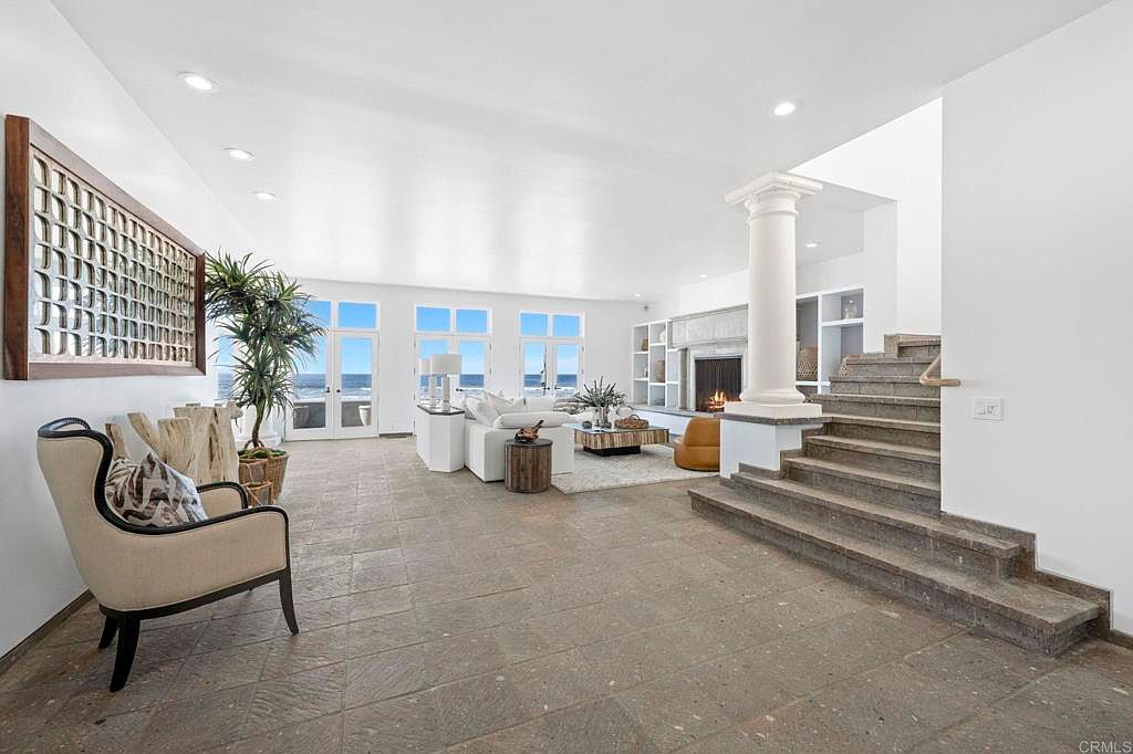 This is a spacious and bright living room featuring a neutral color palette with white walls and light-colored tile flooring. The room has large windows offering ocean views, a fireplace, and a staircase leading to another level. The overall impression is luxurious and airy, perfect for a high-end real estate listing.