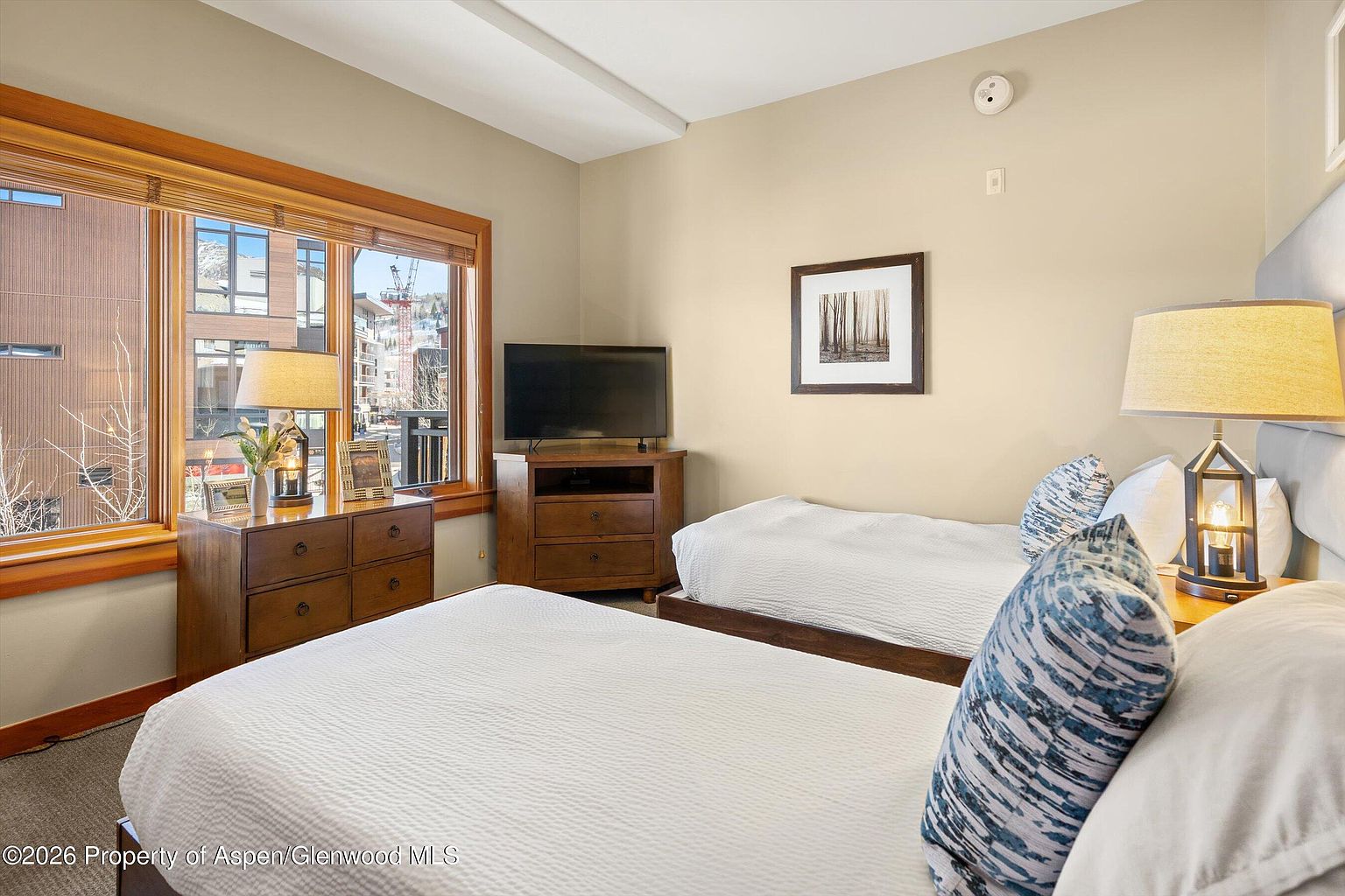 This bedroom features two twin beds with white linens and decorative blue pillows. A wooden dresser sits beneath a window, and a TV is placed on a matching wooden stand. The walls are painted in a neutral tone, and a framed picture hangs above one of the beds, creating a cozy and inviting atmosphere.