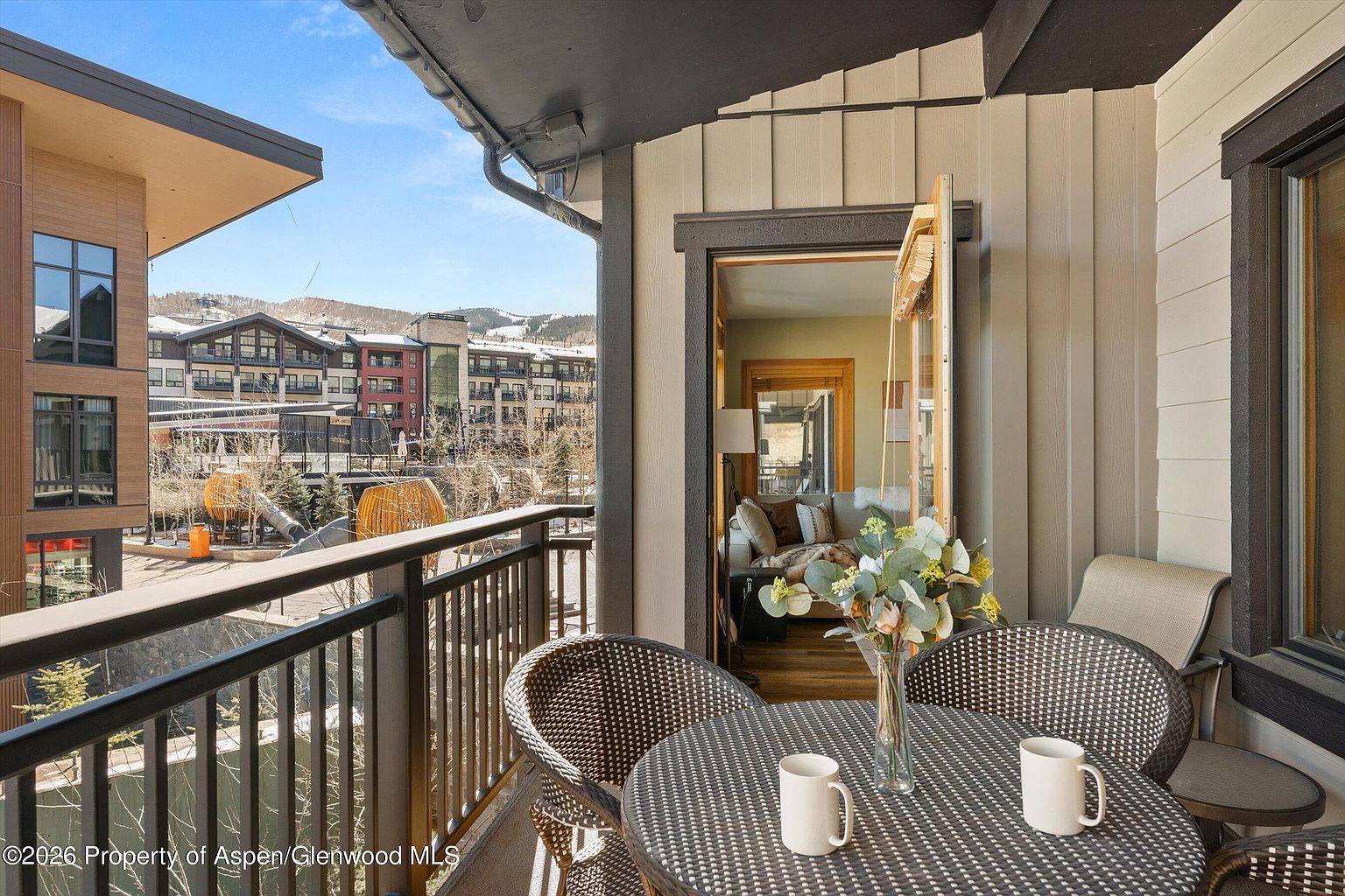 This image showcases a charming balcony setting with a round table and wicker chairs, complete with mugs, suggesting a cozy spot for relaxation. The balcony overlooks a scenic view of buildings and distant mountains, framed by a black railing. An open doorway leads into an interior space, hinting at a seamless indoor-outdoor living experience.