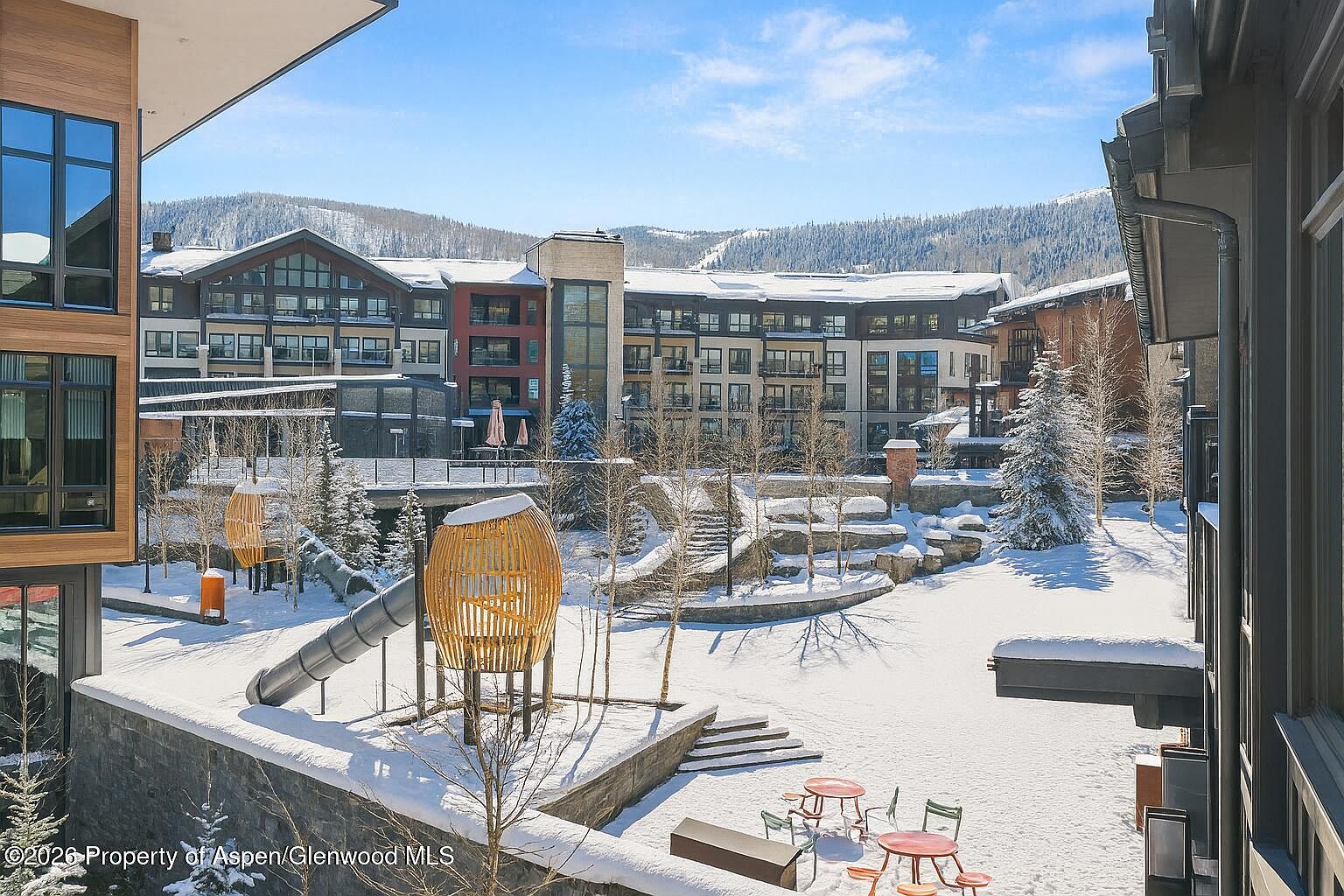 This image showcases a snow-covered community amenity area, possibly a park or recreational space, featuring modern buildings in the background. A playground with a slide and unique wooden structures is visible, along with outdoor seating areas covered in snow. The scene presents a serene winter landscape, suggesting a family-friendly environment within the community.