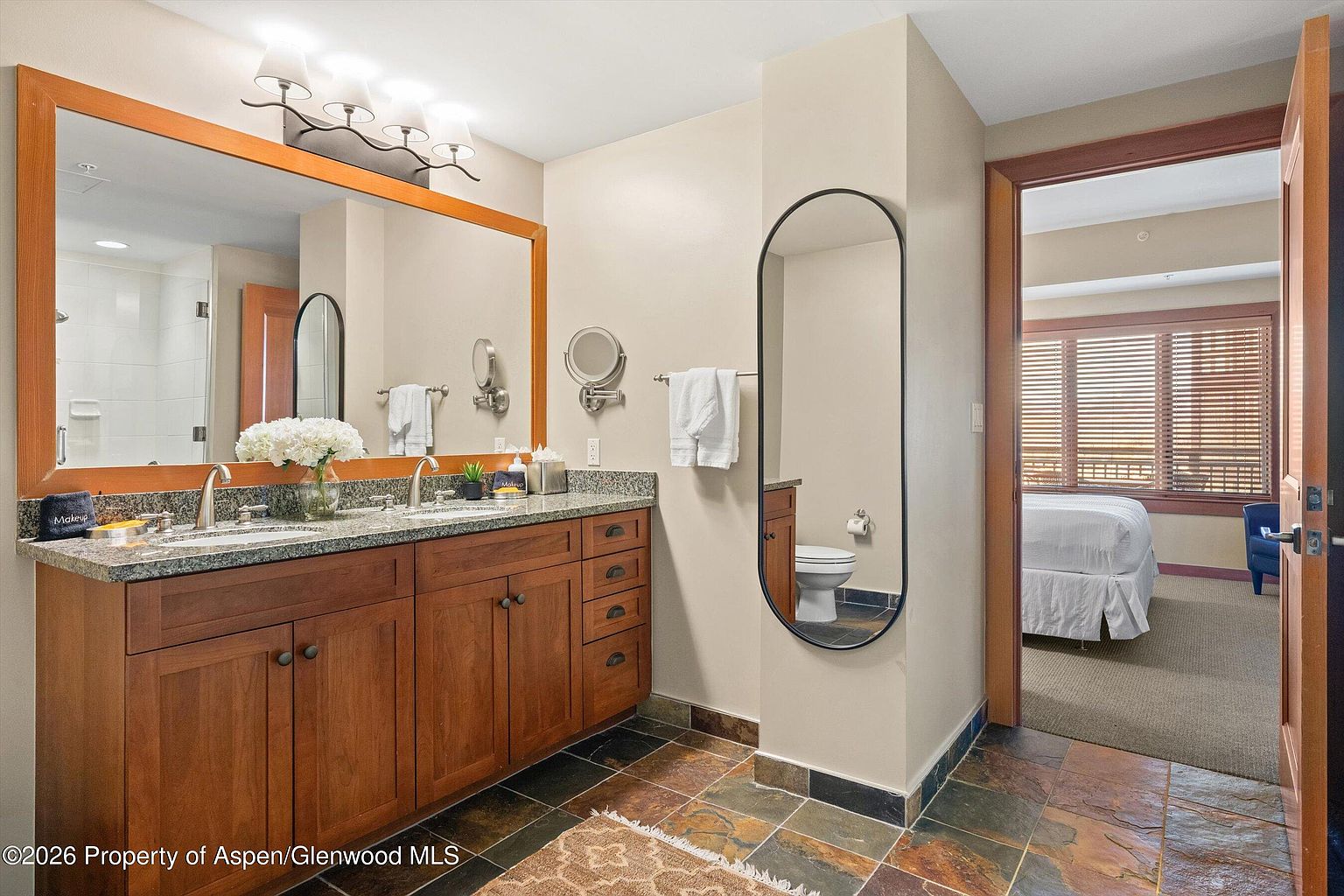 This is a well-appointed primary bathroom featuring a double vanity with a granite countertop and wooden cabinetry. A large mirror hangs above the vanity, reflecting the bright lighting. The bathroom also includes a tall, narrow mirror and a toilet, with a glimpse into an adjacent bedroom through an open doorway, showcasing a bed and window with blinds.