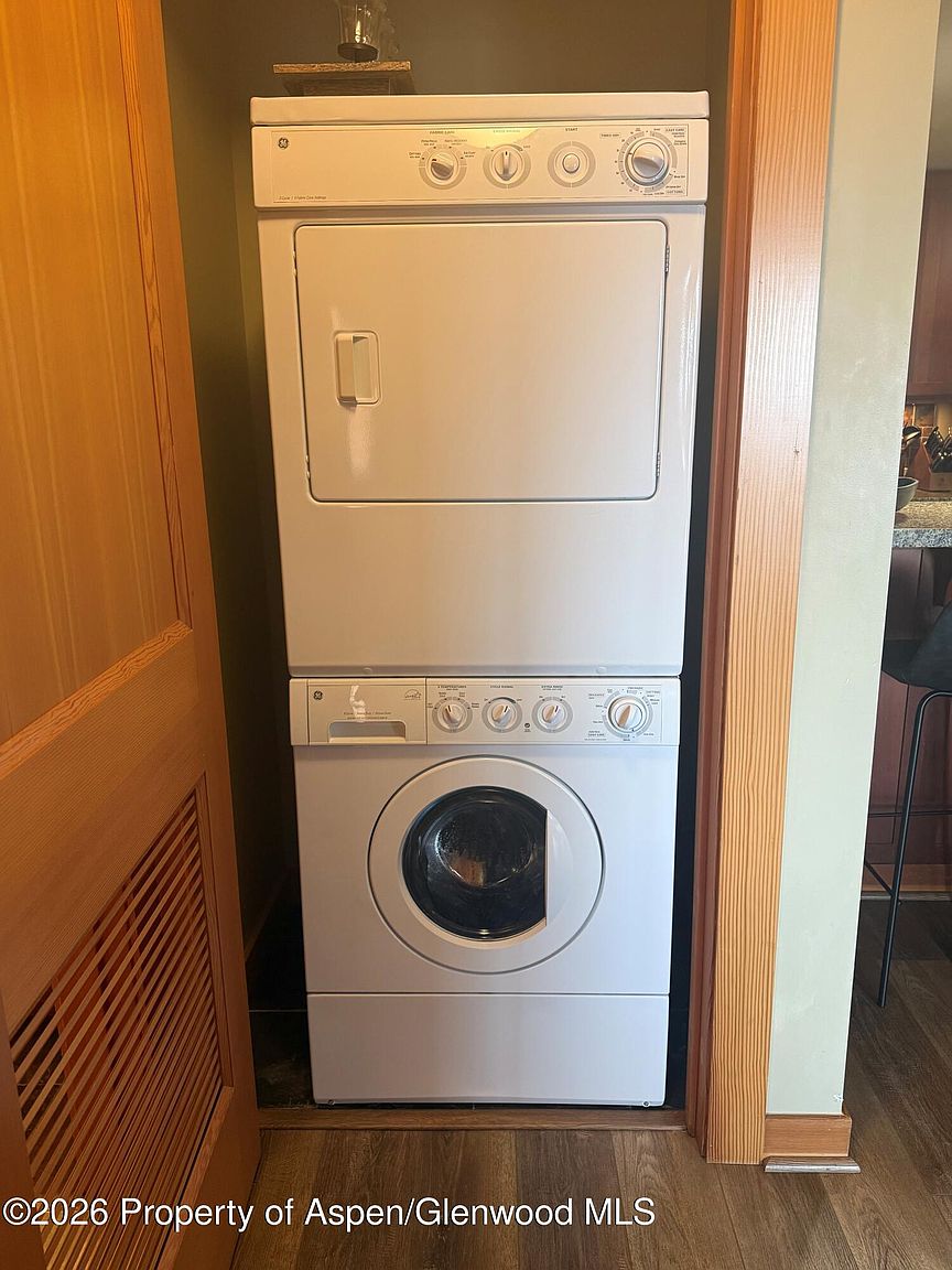 The image showcases a stacked washer and dryer unit, likely within a laundry room or closet. The appliances are white and appear to be in good condition. The surrounding area includes wooden trim and a partially visible door, suggesting a compact and functional space.