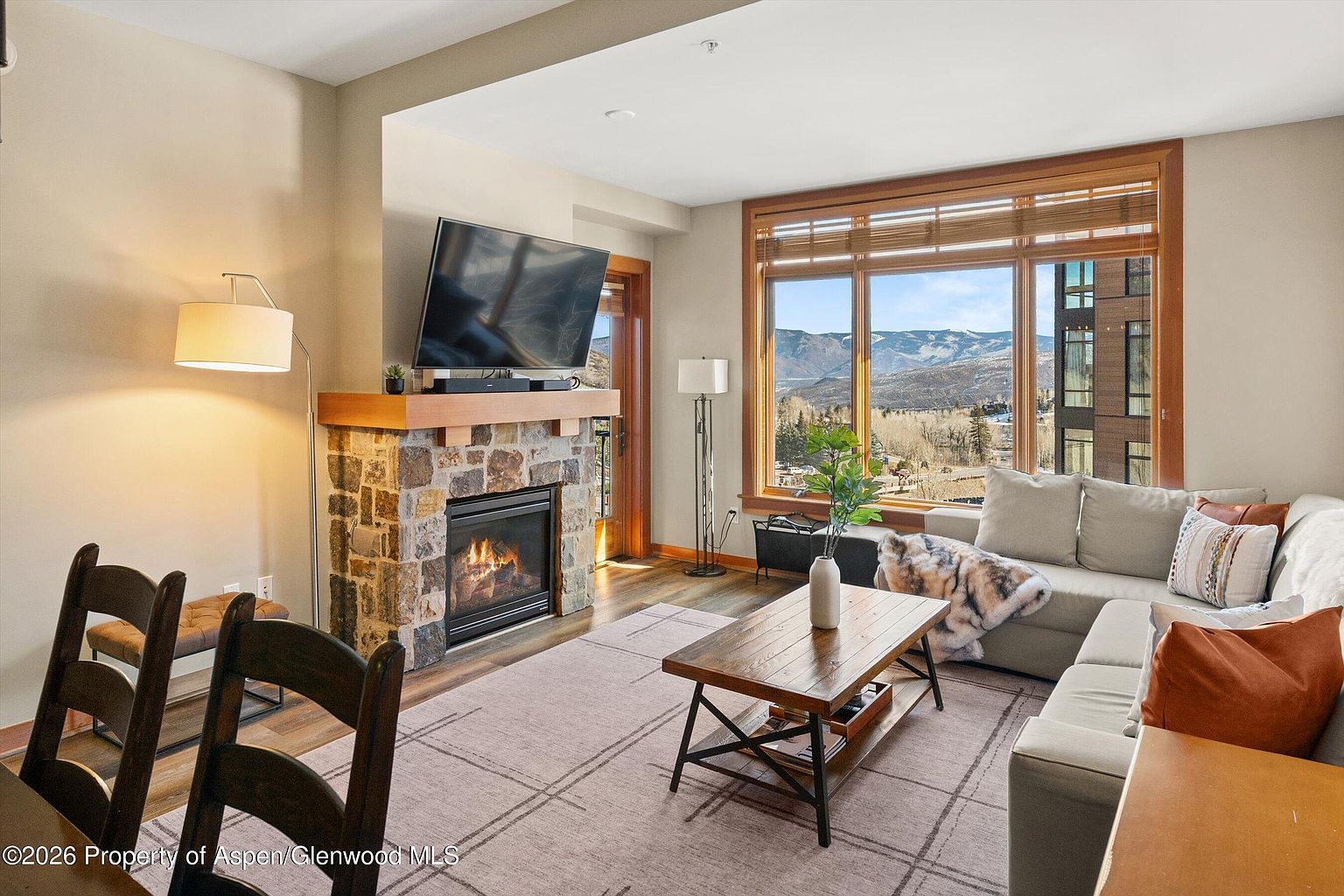 This is an interior shot of a living room featuring a stone fireplace with a mounted television above it. A large window offers a scenic view of mountains and a town, complemented by a comfortable sectional sofa and a wooden coffee table. The room is well-lit and appears cozy, with a neutral color palette and a mix of textures.