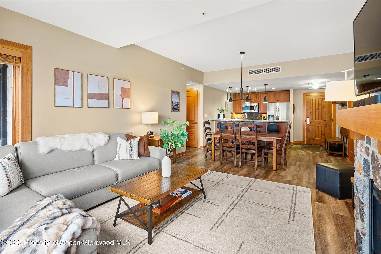 This interior shot showcases a combined living and dining area, with a cozy living room featuring a gray sofa, a wooden coffee table, and artwork on the wall. The dining area includes a wooden table with chairs, adjacent to an open kitchen with wooden cabinetry and a breakfast bar. The space is well-lit and appears inviting, with a warm color palette and a mix of textures.