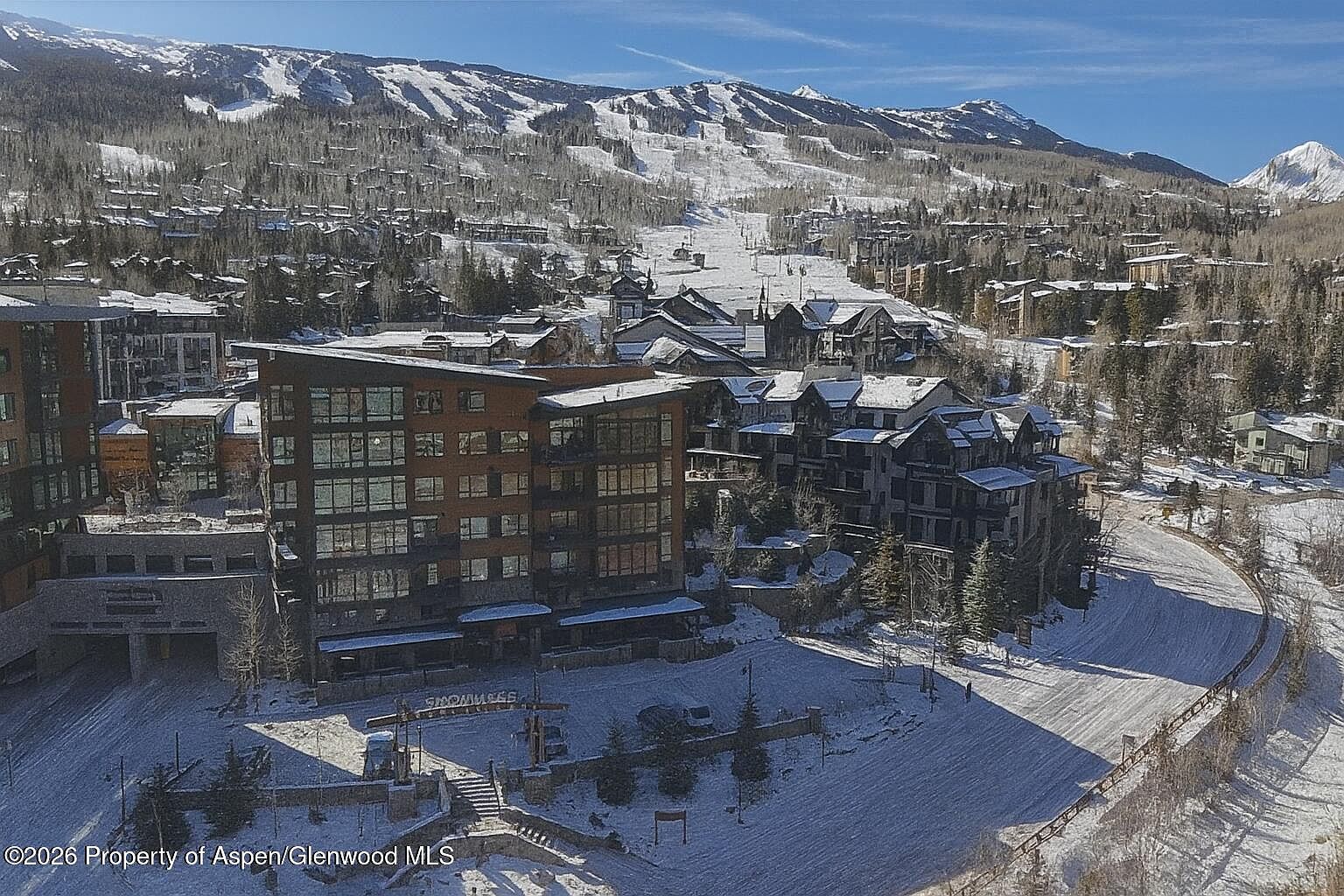This aerial view showcases a luxurious ski resort property nestled in a snowy mountain landscape. Modern architecture blends with traditional chalet styles, featuring multi-story buildings with large windows and snow-covered roofs. The surrounding area includes ski slopes and evergreen trees, creating a picturesque and inviting atmosphere for potential buyers seeking a winter retreat.