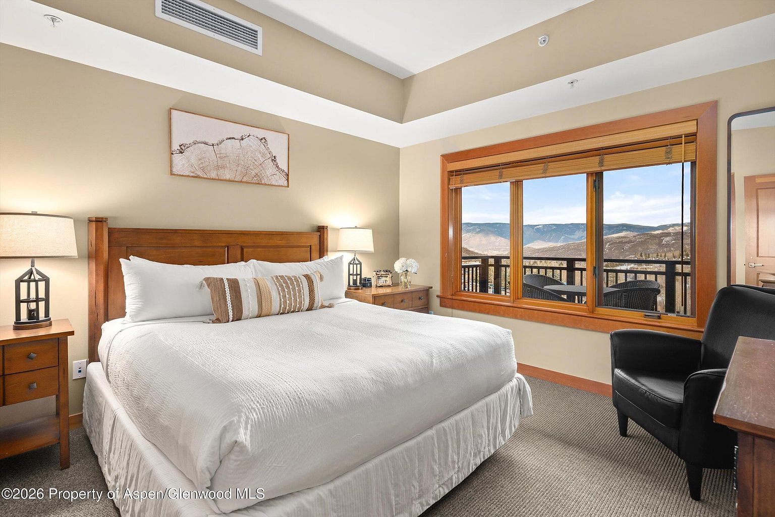 This is a well-lit primary bedroom featuring a king-size bed with white linens and a decorative pillow. The room has a wooden headboard, matching nightstands with lamps, and a large window offering a scenic mountain view. An armchair sits near the window, creating a cozy reading nook.