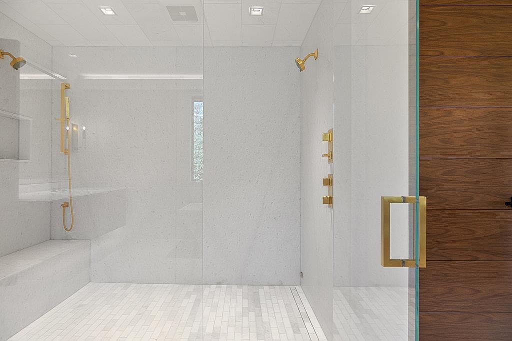 This is a luxurious primary bathroom featuring a spacious walk-in shower with glass enclosure, gold fixtures, and a built-in bench. The walls and floor are tiled in a light color, creating a bright and clean aesthetic. A modern wooden door adds a touch of warmth and sophistication to the space.