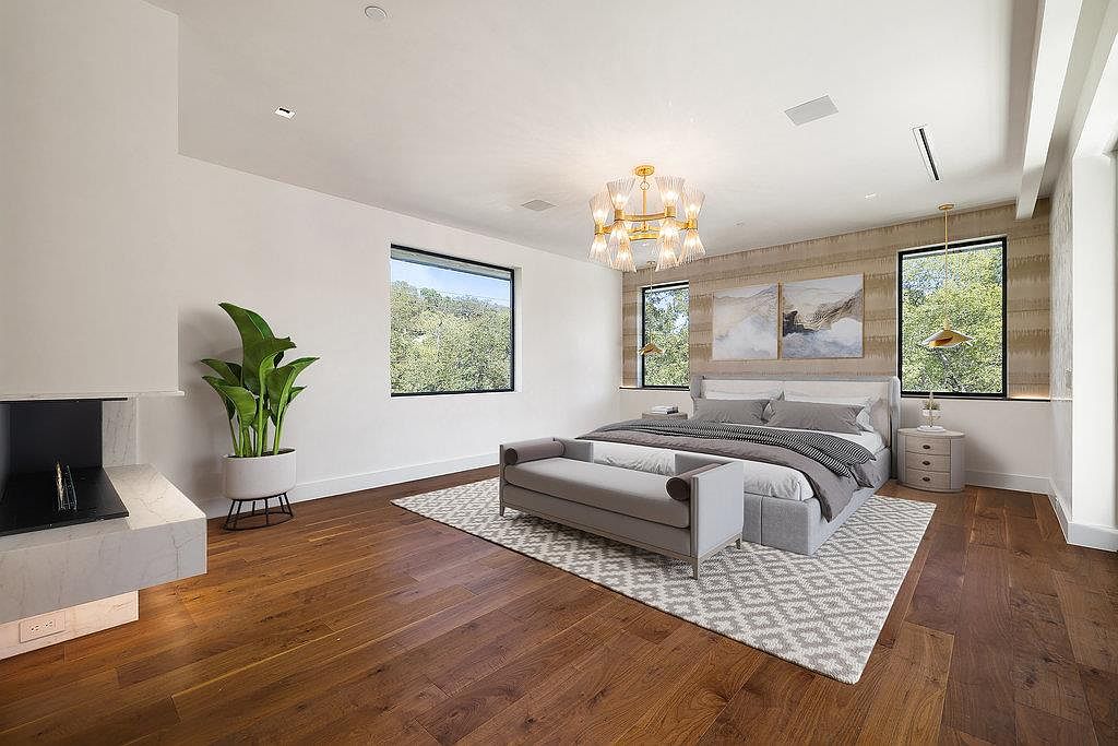 This is a primary bedroom featuring hardwood floors, a large bed with gray bedding, and a gray bench at the foot of the bed. The room has a modern chandelier, two windows providing natural light, and a fireplace on the left. The overall impression is luxurious and serene.