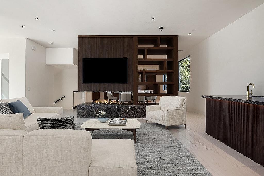 This is a well-lit living room featuring a modern fireplace with a dark marble surround and a large flat-screen TV mounted above. The room includes a comfortable sectional sofa, an accent chair, and a stylish coffee table on a gray rug. The open floor plan and neutral color palette create a sophisticated and inviting atmosphere.