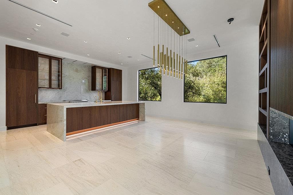 This is a spacious, modern kitchen featuring dark wood cabinetry and a large island with a light-colored countertop. A striking gold pendant light fixture hangs above the island, and two large windows provide natural light and views of the greenery outside. The light-colored tile flooring adds to the bright and airy feel of the space.