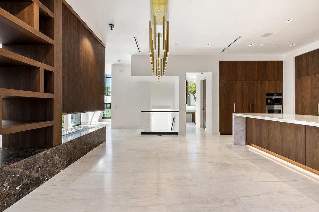 This interior shot showcases a spacious living area with a modern aesthetic. The room features a striking fireplace with a dark marble base and a built-in wooden bookshelf, complemented by a contemporary chandelier. The open floor plan and light-colored tile flooring contribute to a bright and airy atmosphere, ideal for entertaining or relaxing.