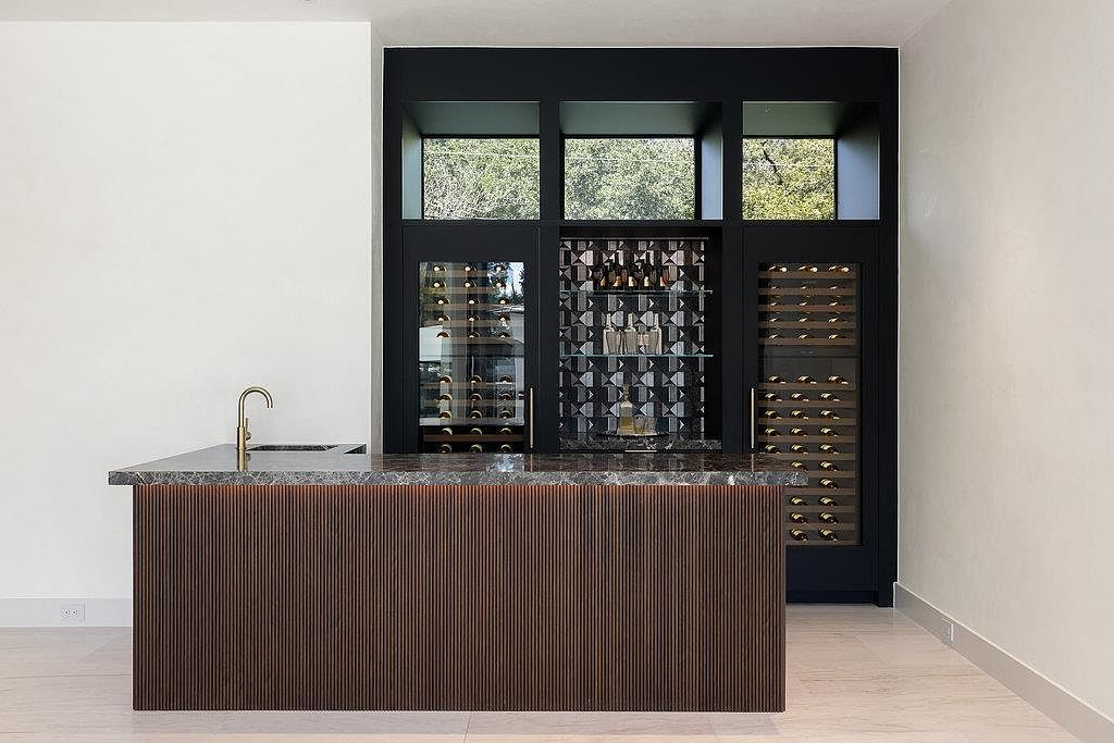 This image showcases a modern wine cellar featuring a dark-toned bar with a textured front and a marble countertop, complemented by a gold faucet. Behind the bar, a built-in wine storage unit with glass doors and open shelving displays numerous wine bottles, set against a backdrop of black framing and small windows that provide natural light. The overall design exudes sophistication and functionality, perfect for wine enthusiasts.