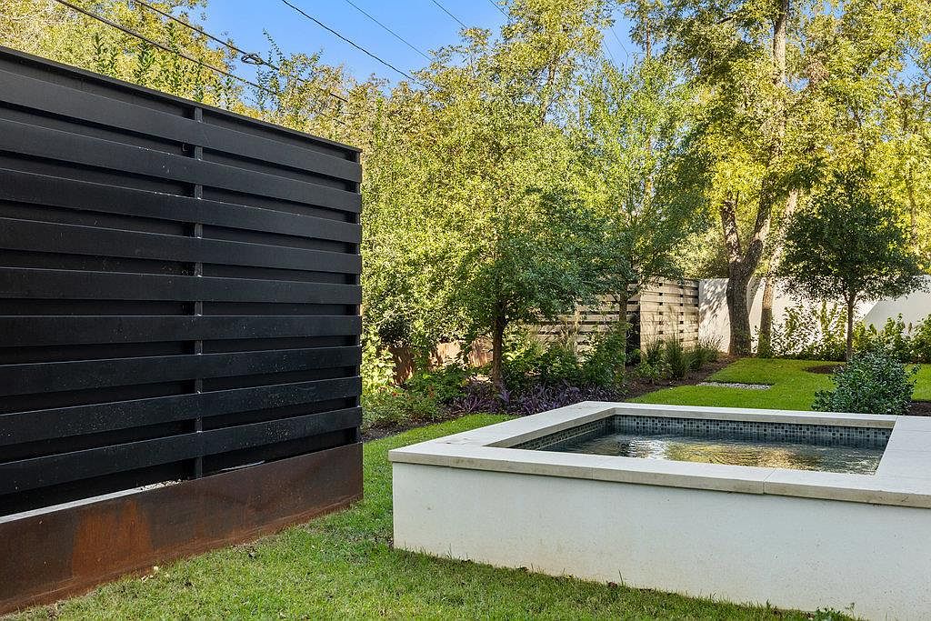 This image showcases a modern backyard garden featuring a small, square pool or spa with a light-colored surround. A dark, horizontal slat fence provides privacy, contrasting with the lush green lawn and mature trees in the background. The overall aesthetic is clean, contemporary, and private, suggesting a relaxing outdoor space.