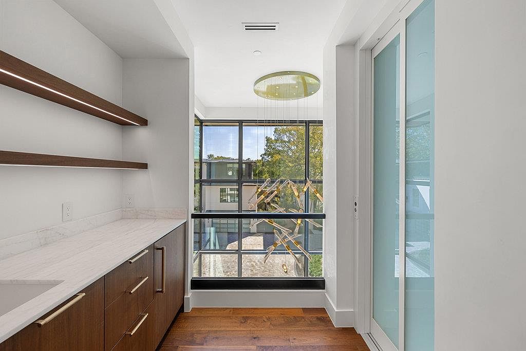 This is a well-lit pantry featuring custom cabinetry with sleek, modern hardware and a light countertop. Open shelving with integrated lighting adds a touch of elegance, while a large window with a unique chandelier provides natural light and a view to the outside. The hardwood flooring and clean lines contribute to a sophisticated and organized space.