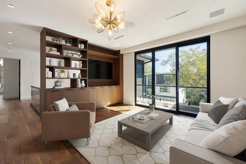 This is an interior shot of a living room featuring a modern design with hardwood floors, a light-colored area rug, and a large window offering natural light. The room includes a built-in shelving unit with a TV, a sofa, an armchair, and a coffee table, creating a comfortable and stylish living space. The overall impression is one of sophistication and comfort.