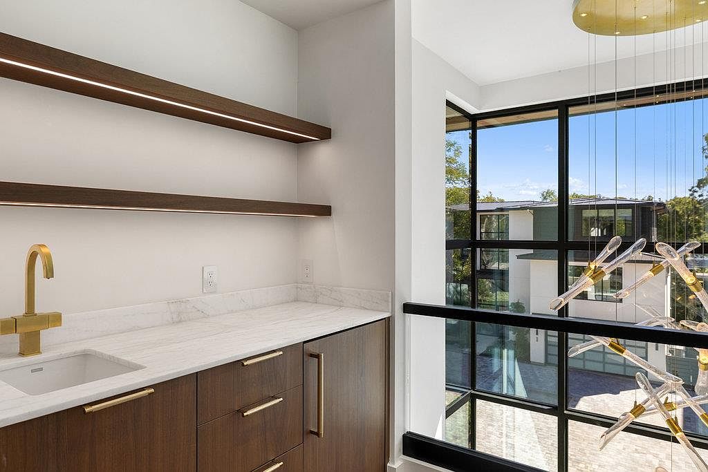 This image showcases a modern pantry area with dark wood cabinetry, gold hardware, and a white marble countertop. Two floating shelves with integrated lighting add a touch of elegance, while a gold faucet and sink provide functionality. A large window offers natural light and a view of the exterior.