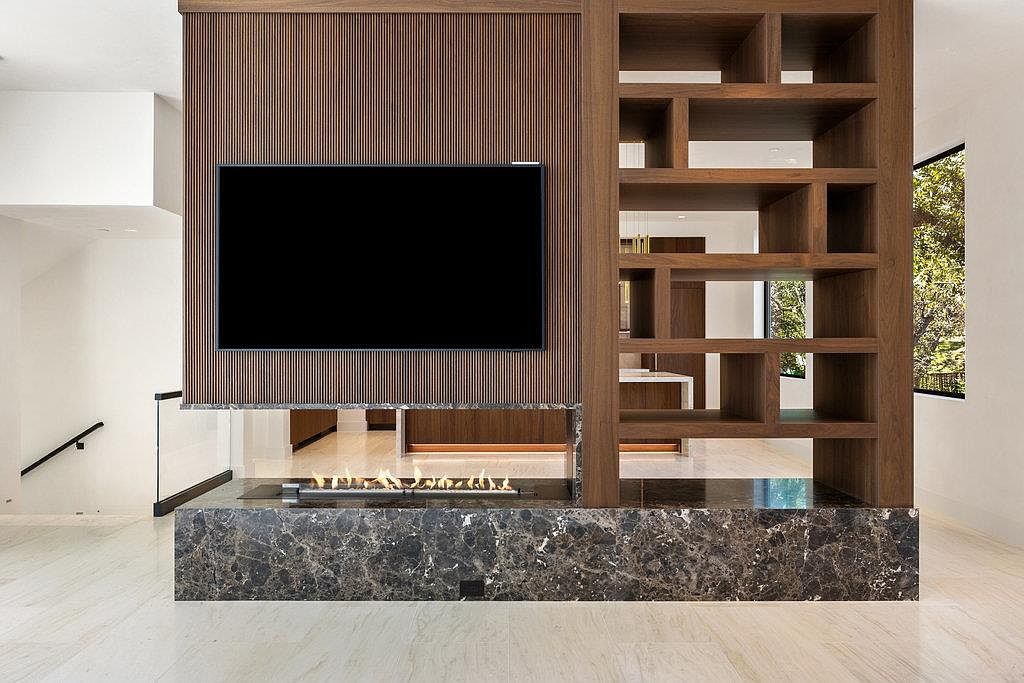 This interior shot showcases a modern living room with a striking architectural feature: a combined media wall and room divider. The wall incorporates a large flat-screen TV mounted on a wood-paneled section, complemented by an open shelving unit and a sleek, linear fireplace with a dark marble base. The design creates a sophisticated and inviting atmosphere.