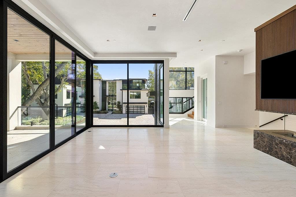 This is a spacious, modern living room with large windows offering an expansive view of the exterior. The room features light-colored tile flooring, a minimalist fireplace, and a sleek, contemporary design. The open floor plan and abundant natural light create an airy and inviting atmosphere.