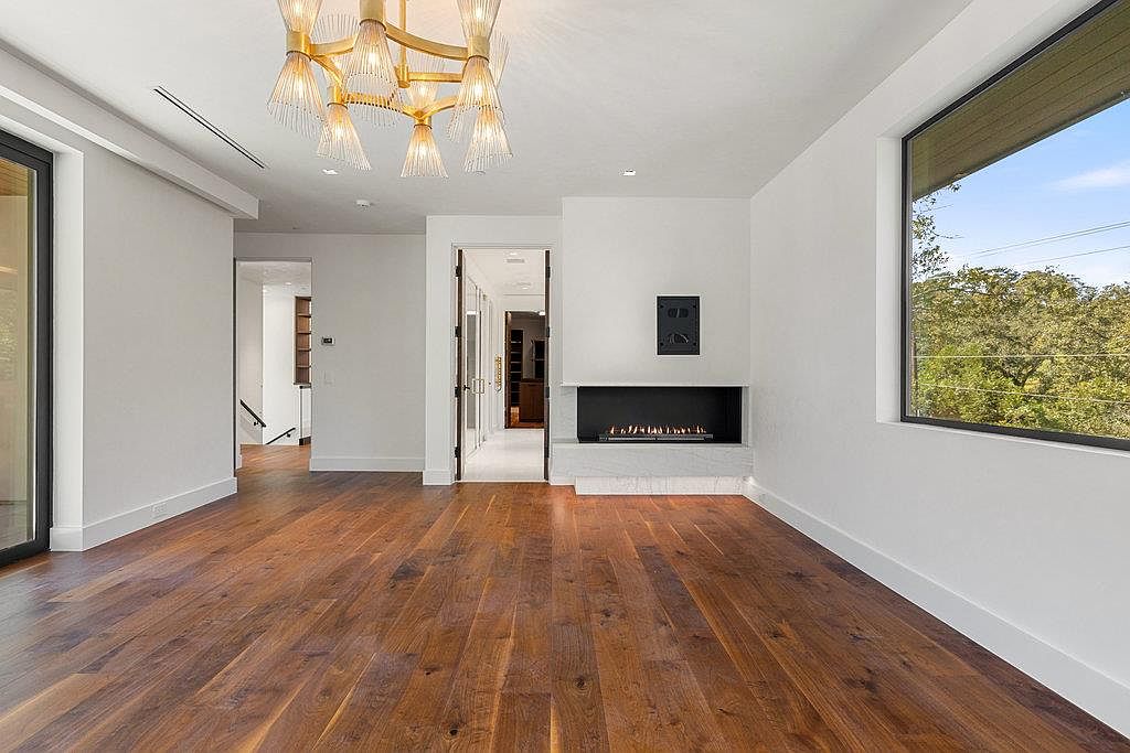 This is an interior shot of a modern living room featuring hardwood floors and white walls. A contemporary fireplace is centered on one wall, and a large window provides natural light. An ornate chandelier hangs from the ceiling, adding a touch of elegance to the minimalist design.