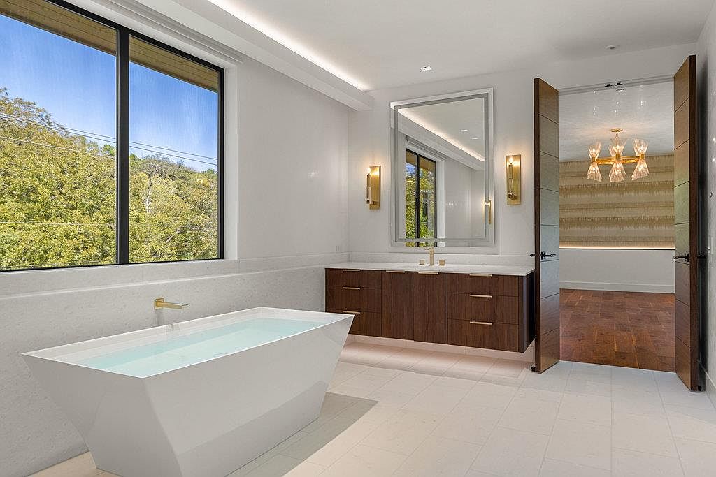 This is a luxurious primary bathroom featuring a modern freestanding bathtub filled with water, a floating vanity with dark wood cabinets and a light countertop, and a large mirror with sconces. The room is bright and airy, with a large window offering natural light and a view of the trees outside. An open doorway leads to another room, showcasing hardwood floors and a decorative chandelier.