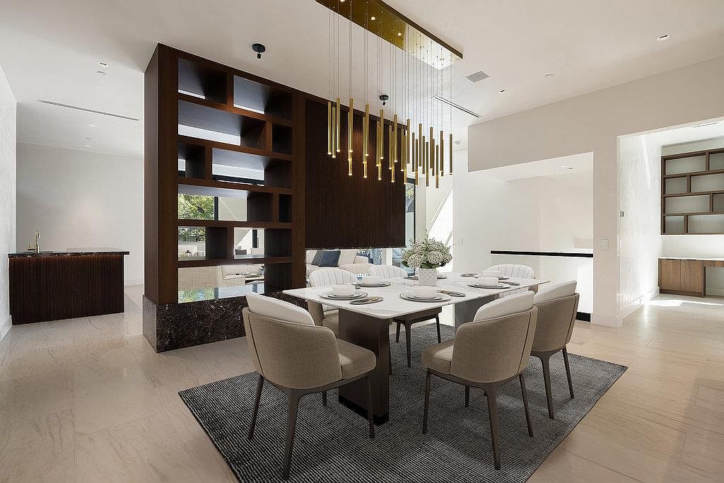 This is an interior shot of a modern dining room. The room features a rectangular dining table with seating for six, a unique chandelier with hanging gold rods, and a dark wood shelving unit that partially divides the space. The color palette is neutral with beige chairs and a gray rug, creating a sophisticated and inviting atmosphere.