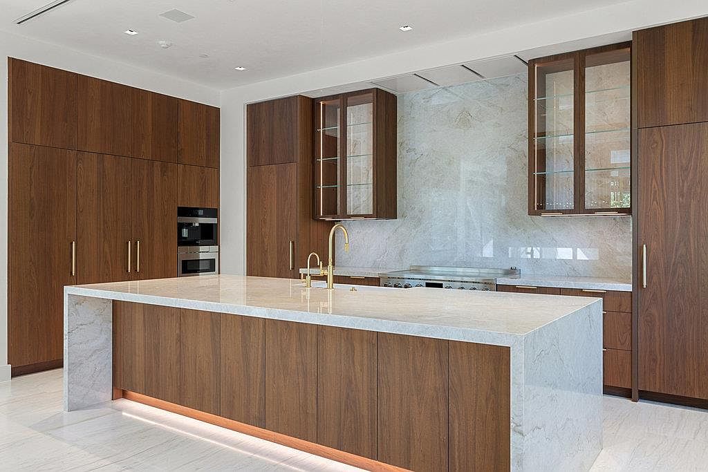 This is a modern kitchen with sleek, dark wood cabinetry and a large island featuring a white marble countertop. The kitchen is well-lit, with a minimalist design and high-end appliances. The overall impression is one of luxury and sophistication.