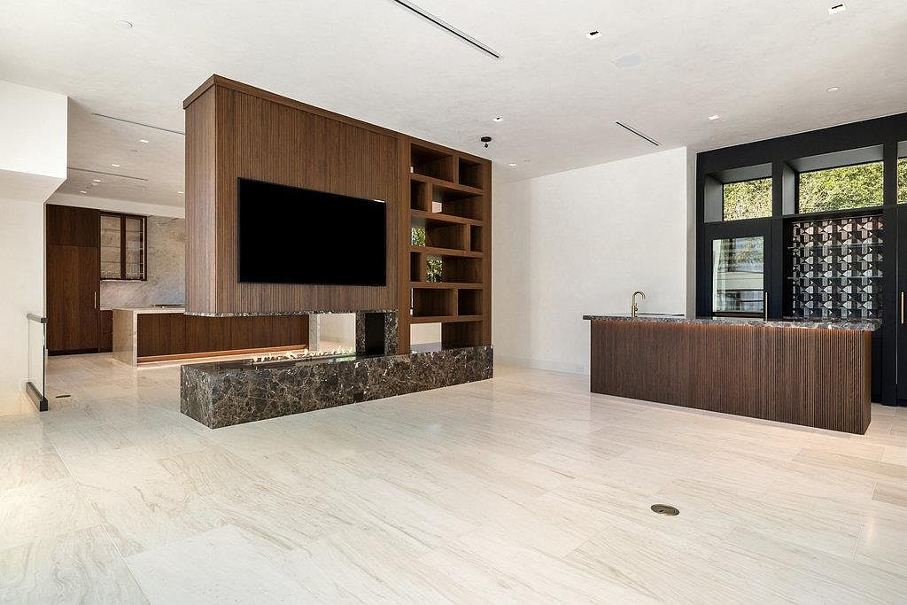 This interior shot showcases a modern living room with a unique architectural design. A central wooden structure houses a large television and open shelving, complemented by a marble-like base with a fireplace. The room features light-colored flooring and a bar area with dark wood cabinetry, creating a sophisticated and spacious atmosphere.