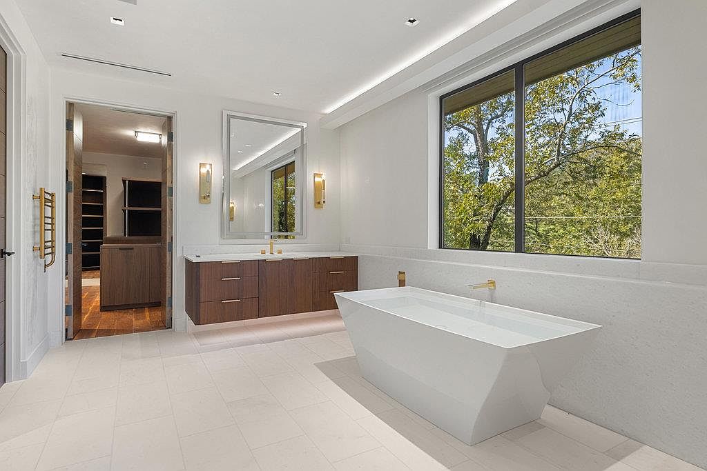 This is a luxurious primary bathroom featuring a modern freestanding bathtub filled with water, a sleek vanity with dark wood cabinetry and gold fixtures, and a large mirror flanked by sconces. The room is bright and airy, with a large window offering a view of the trees outside. The overall impression is one of spa-like tranquility and high-end design.