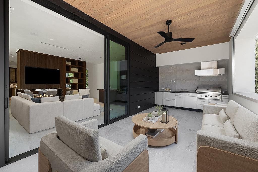 This image showcases a modern outdoor living space featuring a covered patio with a built-in kitchen and comfortable seating. The kitchen area includes stainless steel appliances and sleek cabinetry, while the seating area offers a relaxing space with plush sofas and a coffee table. Large sliding glass doors seamlessly connect the indoor and outdoor spaces, creating an inviting and functional area for entertaining or relaxing.
