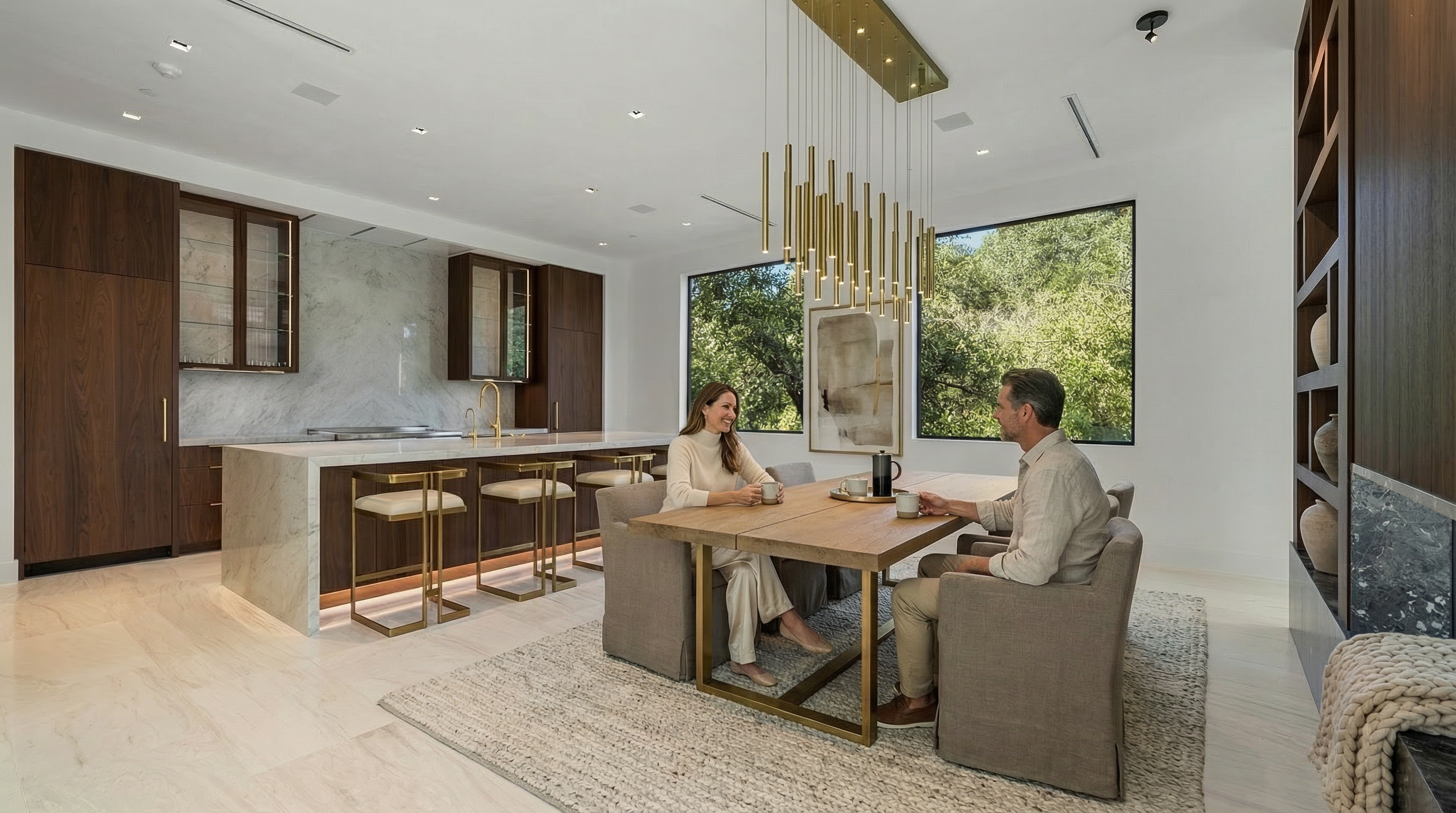 This sophisticated, open-concept kitchen seamlessly integrates a dining area, featuring warm dark wood cabinetry, waterfall marble countertops, and gold-toned accents. A striking tiered pendant light fixture serves as a focal point above the dining table, while large windows provide ample natural light and views of lush greenery. The space exudes a luxury contemporary aesthetic, perfect for both casual meals and refined entertaining.
