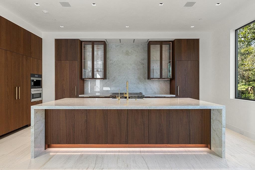 This is a modern kitchen featuring dark wood cabinetry and a large island with a marble countertop. The kitchen is well-lit with recessed lighting and natural light from a nearby window. The overall impression is one of luxury and sophistication, with a focus on clean lines and high-end finishes.