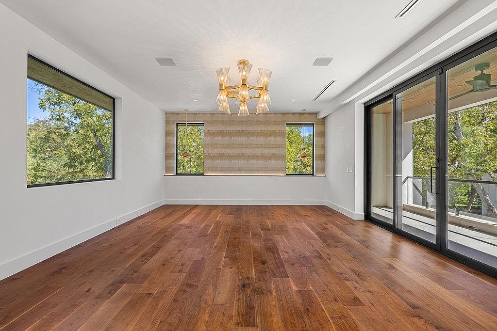 This is a spacious living room featuring hardwood floors, white walls, and large windows that provide ample natural light. A modern chandelier hangs from the ceiling, and sliding glass doors lead to an outdoor area. The room has a clean and contemporary aesthetic, perfect for entertaining or relaxing.