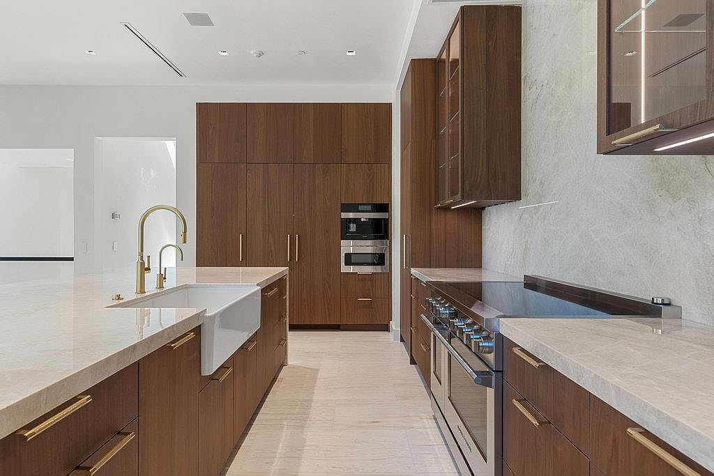 This is a modern kitchen featuring dark wood cabinetry with gold hardware and light countertops. The kitchen includes a large farmhouse sink with a gold faucet, stainless steel appliances, and a built-in oven and microwave. The overall impression is sleek and luxurious.