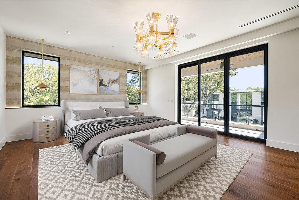 This is a primary bedroom featuring a large bed with gray linens, a bench at the foot of the bed, and a patterned rug. The room has a modern chandelier, two windows, and a large sliding glass door leading to a balcony. The walls are a mix of textured wallpaper and white paint, creating a sophisticated and inviting space.