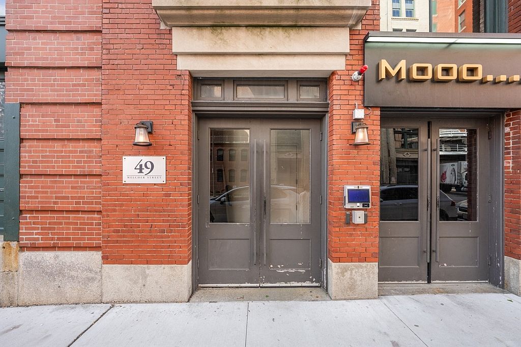 This image showcases the exterior entryway of a brick building, featuring two adjacent doorways. The left entrance is marked with a sign reading '49 Melcher Street' and is flanked by a vintage-style lantern. To the right, a second entrance is identified by a 'Mooo...' sign, suggesting a restaurant or business, and is also illuminated by a similar lantern, creating a welcoming and classic aesthetic.