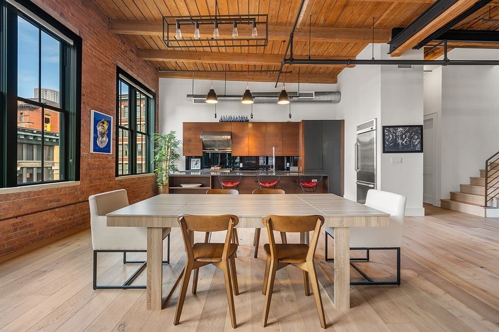 This interior shot showcases a dining area adjacent to a modern kitchen. The dining space features a light wood table surrounded by a mix of wooden and upholstered chairs, set against a backdrop of exposed brick and large windows. The kitchen area includes sleek cabinetry and stainless steel appliances, creating a stylish and inviting atmosphere.