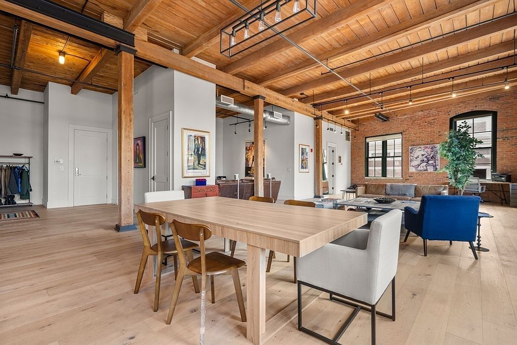 This interior shot showcases a spacious dining area within a modern loft. The room features a large wooden dining table surrounded by chairs, set against a backdrop of exposed brick walls, wooden beams, and hardwood floors. Natural light streams in through the windows, highlighting the open-concept design and industrial-chic aesthetic.