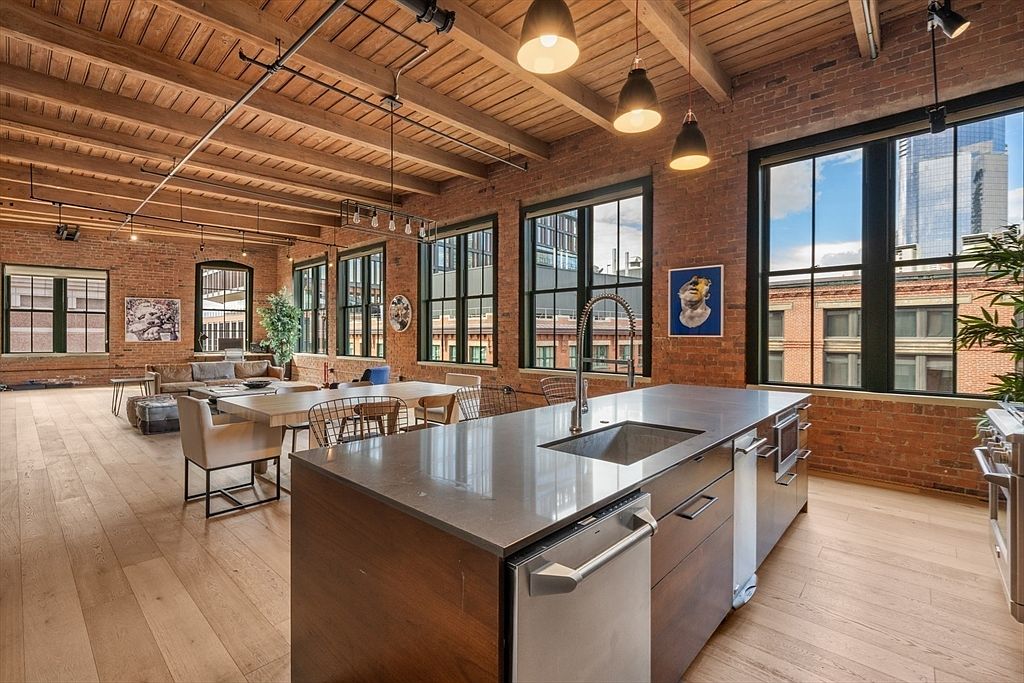 This interior shot showcases a modern kitchen in a loft-style apartment, featuring a large island with a sink, stainless steel appliances, and wooden cabinetry. The kitchen is open to a dining area with a large wooden table and chairs. Exposed brick walls, large windows with city views, and wooden ceilings with industrial lighting fixtures contribute to the space's urban and contemporary aesthetic.