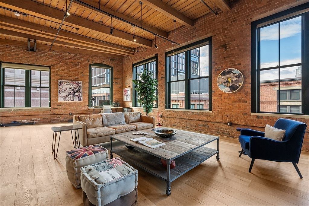 This is an interior shot of a living room featuring exposed brick walls, large windows, and wooden ceilings with exposed beams. The room is furnished with a leather sofa, a blue armchair, a rustic wooden coffee table, and patterned ottomans, creating a blend of industrial and contemporary styles. The space is well-lit and appears spacious, highlighting the architectural details and comfortable living area.