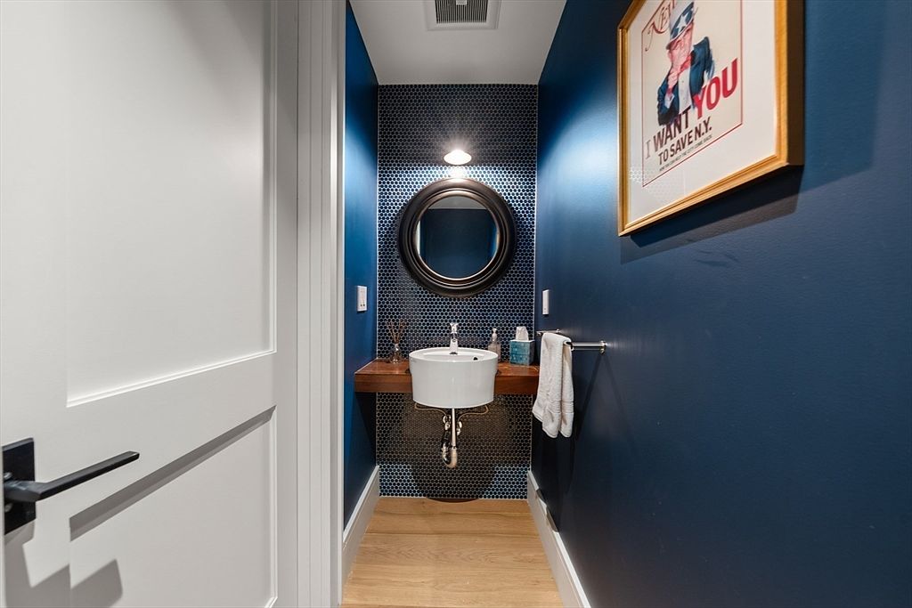 This is a stylish guest bathroom featuring a dark blue color scheme with a round mirror and a wall-mounted sink. The walls are a combination of dark blue paint and small, dark blue mosaic tiles. A framed poster adds a touch of character, and the light wood flooring provides a warm contrast to the cool tones.