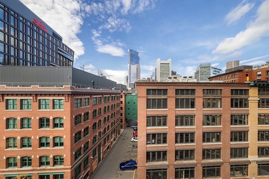 This aerial view showcases a dense urban landscape with brick buildings and modern skyscrapers under a partly cloudy sky. The composition highlights the architectural diversity and the proximity of older structures to contemporary high-rises, creating a dynamic visual contrast. A narrow street with parked cars adds a sense of scale and activity to the scene.