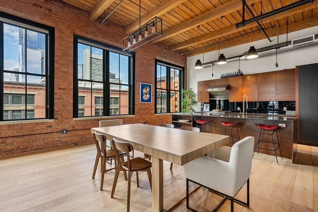This interior shot showcases a dining area adjacent to a modern kitchen. The dining space features a large wooden table surrounded by chairs, with exposed brick walls and large windows offering city views. The kitchen boasts sleek wooden cabinetry and a breakfast bar with stylish stools, creating an open and inviting atmosphere.
