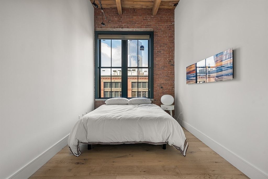 This is an interior shot of a bedroom featuring a bed with white linens, a small side table with a lamp, and a modern art piece on the wall. The room has a minimalist design with white walls, hardwood floors, and a large window offering a view of the city. The exposed brick wall adds a touch of character to the space.