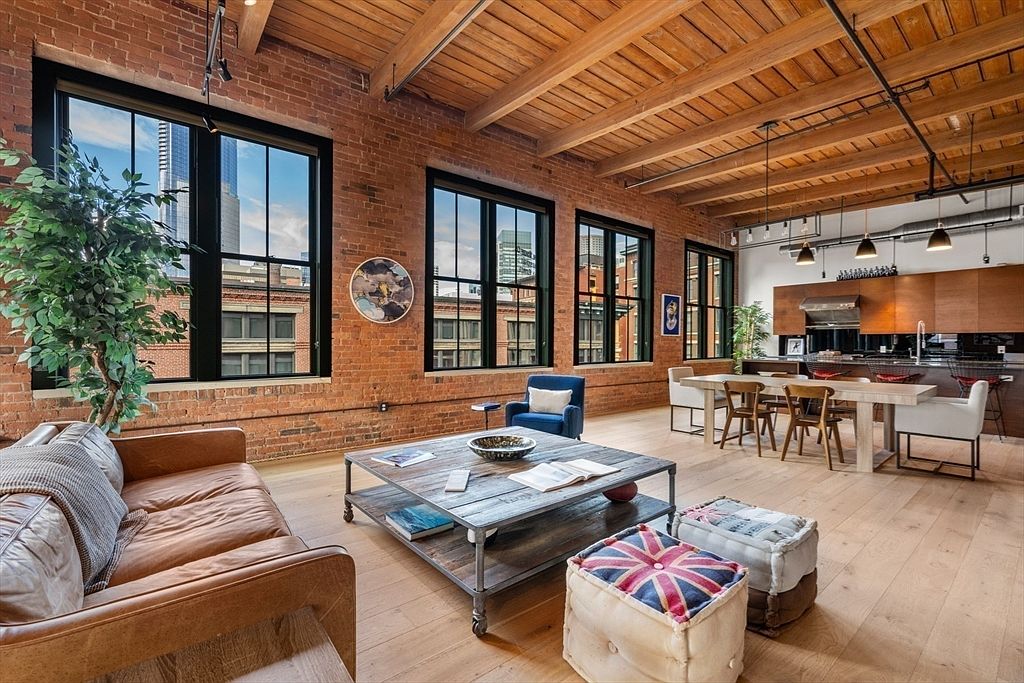This is an interior shot of a spacious living room in a loft-style apartment. The room features exposed brick walls, large windows with black frames, and a wooden ceiling with exposed beams. The furniture includes a leather sofa, a rustic coffee table, and unique ottomans, creating a blend of industrial and comfortable living space.