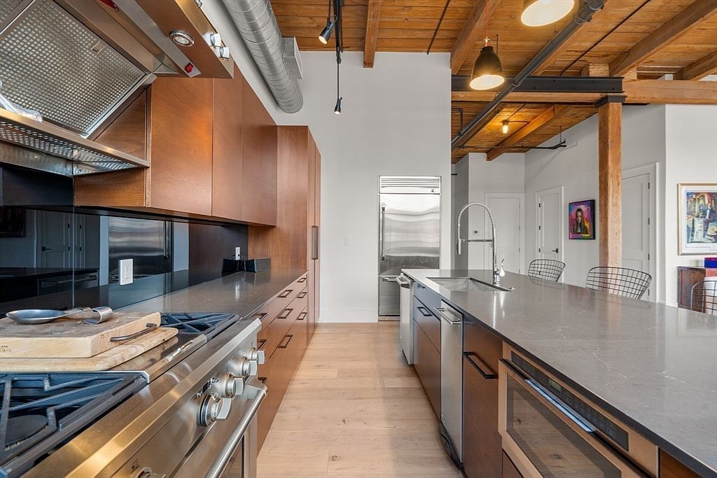 This is a modern kitchen with wooden cabinets and stainless steel appliances. The kitchen features a large island with a gray countertop and a stainless steel sink. The ceiling has exposed wooden beams and industrial-style lighting, adding a unique architectural element to the space.