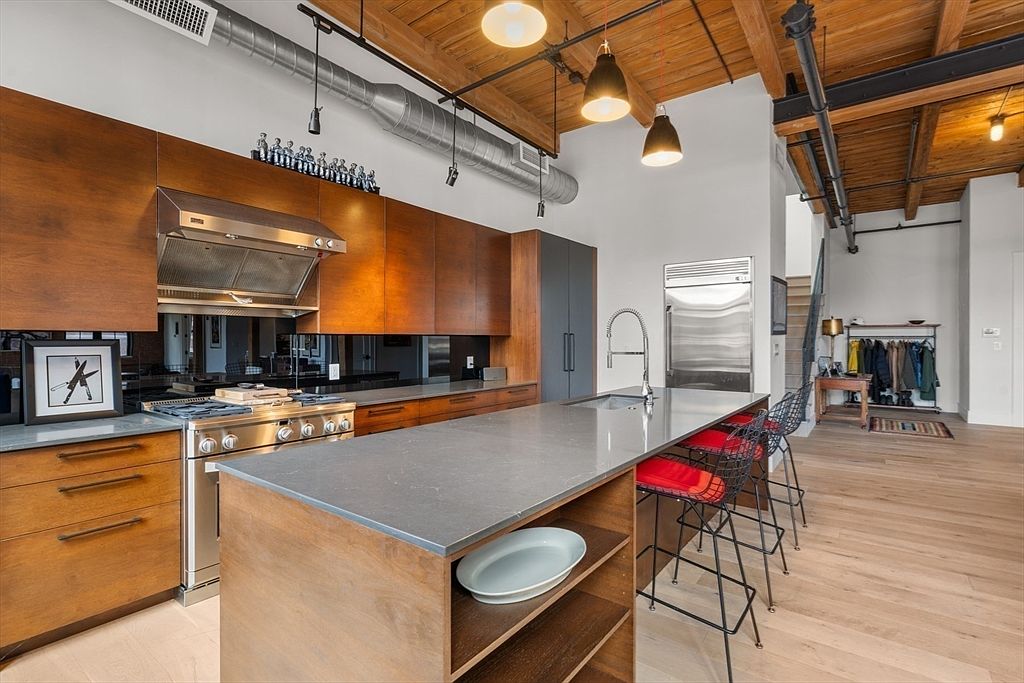 This is a modern kitchen with wooden cabinets, stainless steel appliances, and a large island with a gray countertop. The kitchen features an open layout with exposed wooden beams on the ceiling and industrial-style lighting fixtures. The perspective is from a medium-height angle, showcasing the kitchen's layout and design.