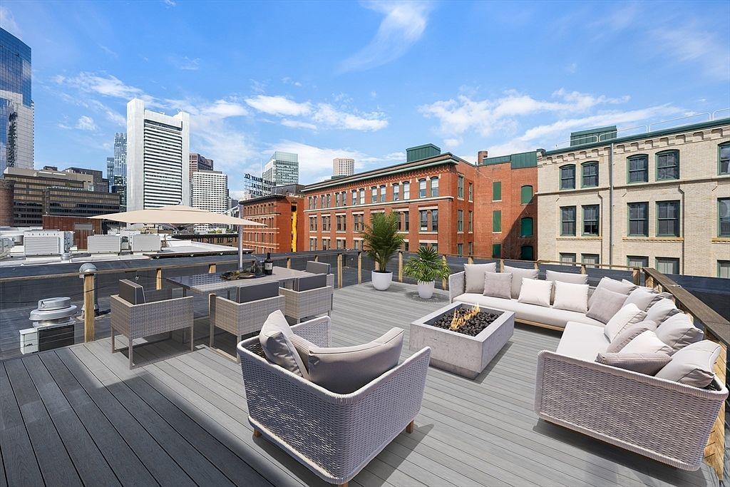 This image showcases a luxurious rooftop patio/deck with stylish outdoor furniture, including a seating area with plush cushions, a dining table set, and a modern fire pit. The deck offers stunning city views, with skyscrapers visible in the background. The overall impression is one of sophisticated urban living and relaxation.