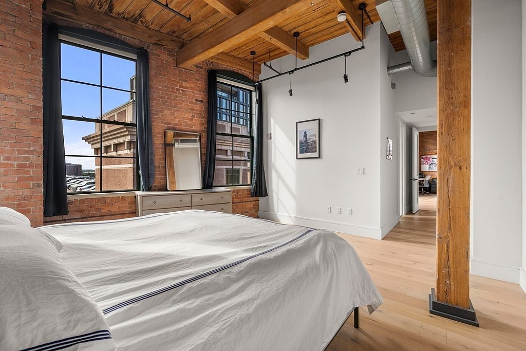 This is a primary bedroom featuring exposed brick walls, large windows with black frames, and a wooden beam ceiling. A white bed with blue stripes is the focal point, complemented by a dresser and a mirror. The room also has a wooden support beam and a doorway leading to another room, creating a loft-like and industrial-chic atmosphere.