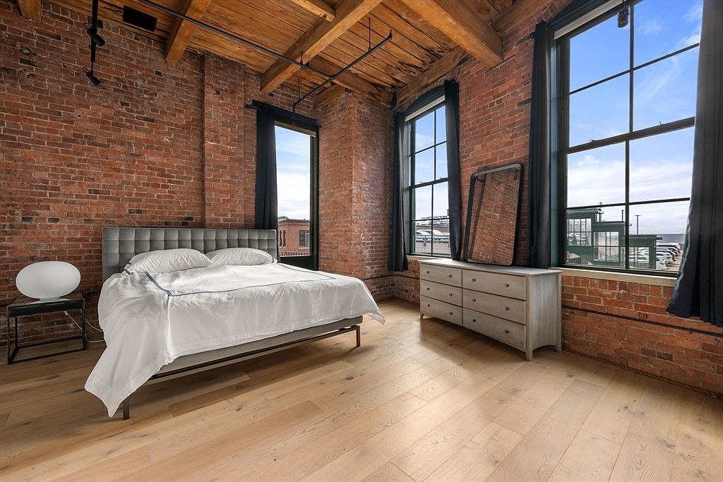 This is a primary bedroom featuring exposed brick walls, wooden beams on the ceiling, and hardwood floors. A large bed with white linens is the focal point, complemented by a dresser and a side table with a modern lamp. Large windows provide natural light and views, enhancing the room's spacious and industrial-chic aesthetic.