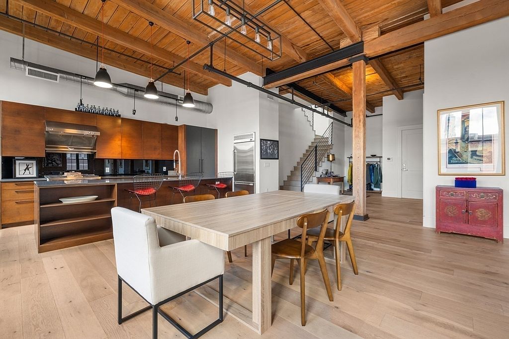 This interior shot showcases a dining area with a large wooden table surrounded by chairs, set against a backdrop of an open-concept living space. The room features hardwood floors, exposed wooden beams on the ceiling, and a modern kitchen area in the background. A staircase is visible, adding to the loft-like feel of the space.